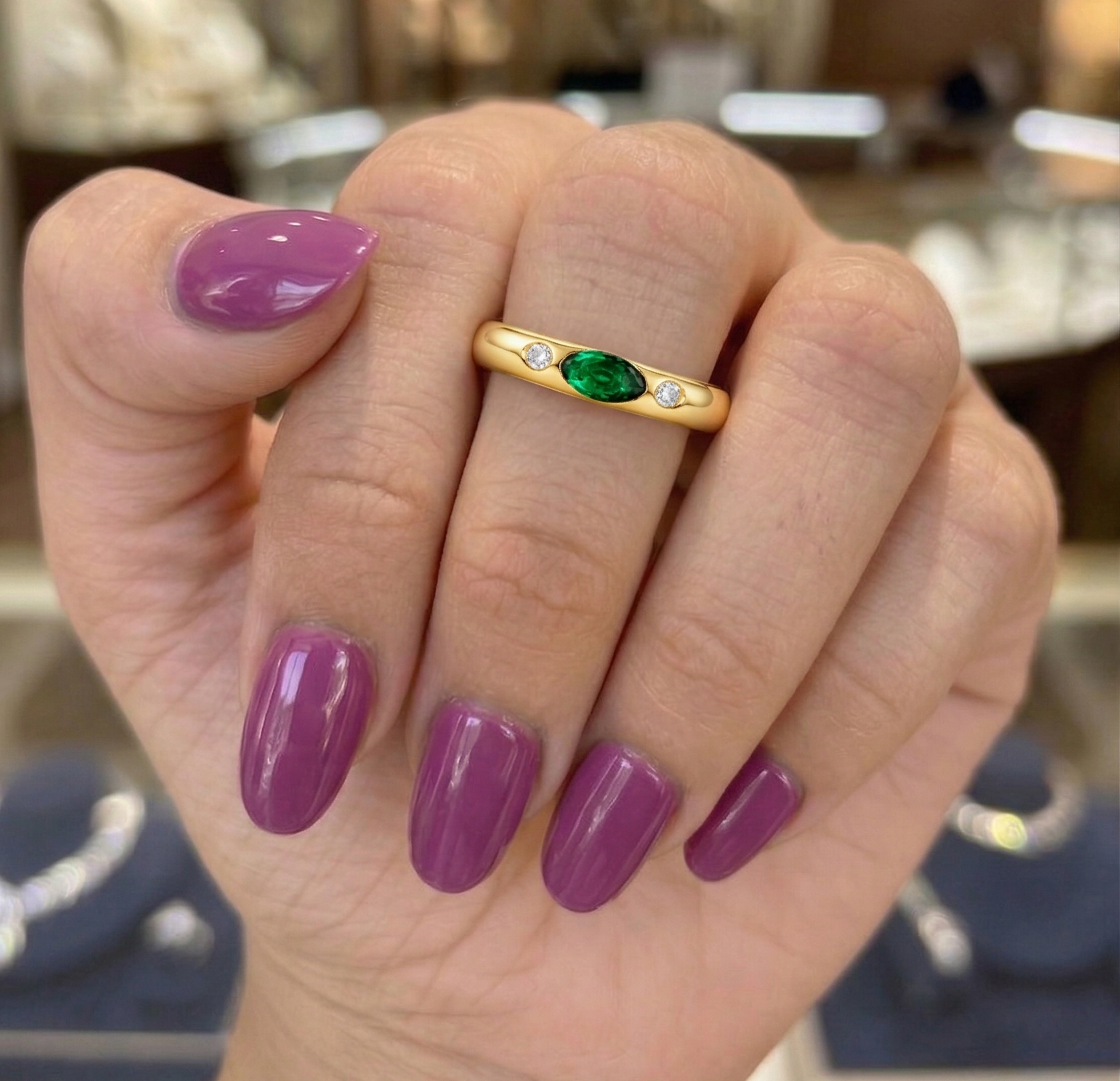 Hand wearing a gold ring with green gemstone in a blurred jewelry store setting