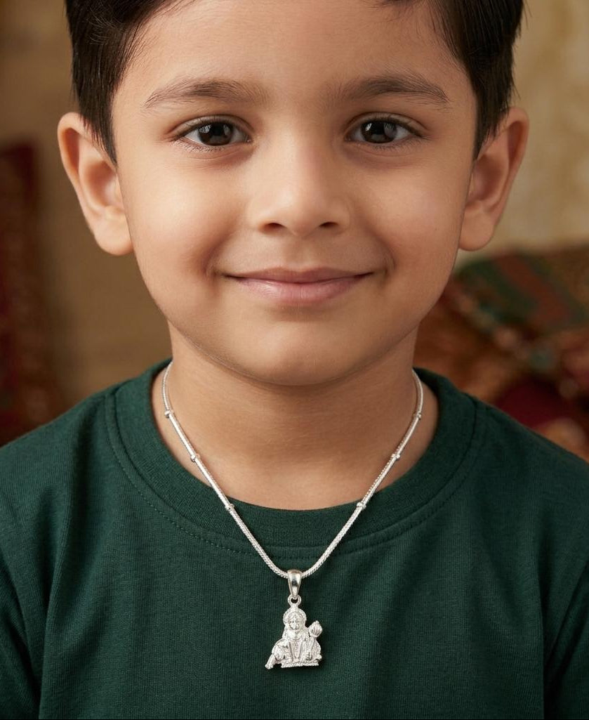 Young boy wearing a green shirt and a necklace with a pendant, sitting in a warm-toned room.