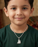Young boy wearing a green shirt and a necklace with a pendant, sitting in a warm-toned room.
